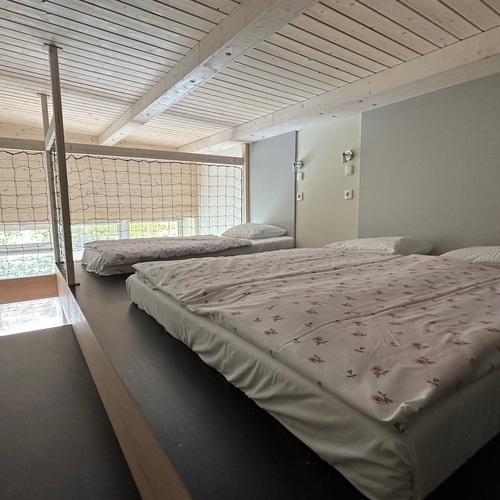 Mezzanine bedroom with two beds, a low whitewashed wooden ceiling, a safety net and bedside lamps.