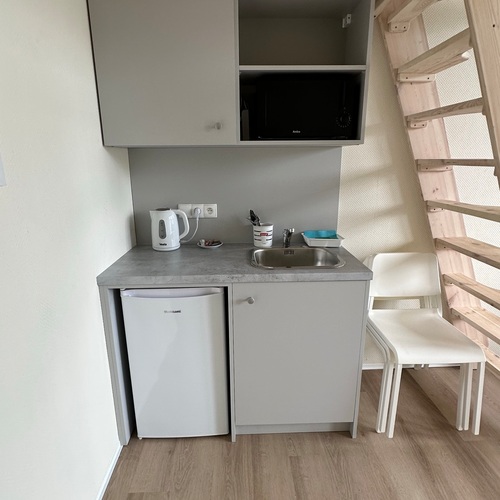 Compact kitchenette with a fridge, sink, kettle, microwave and two chairs by a wooden mezzanine