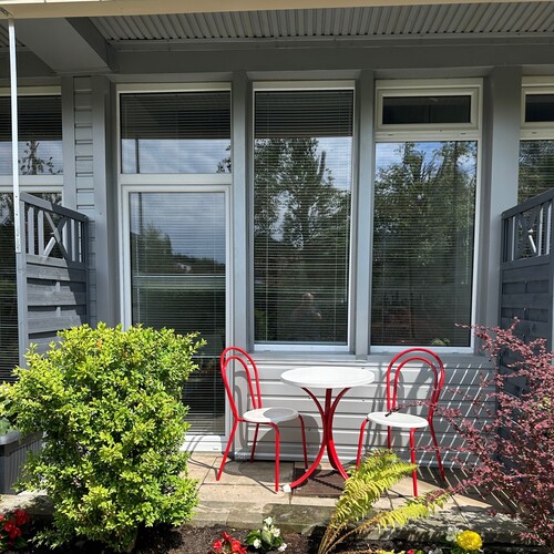 Cozy terrace with a white table and two red bistro chairs in front of large windows, surrounded by greenery and flowers
