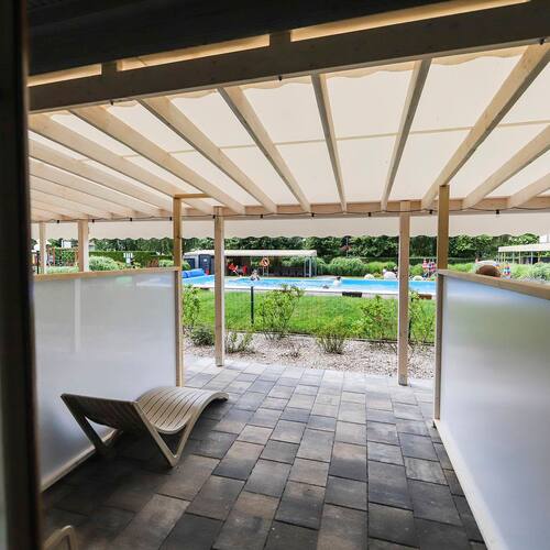 Covered private cabana with a wooden sunbed, paved floor and views of the outdoor pool, lawn and bathing guests.