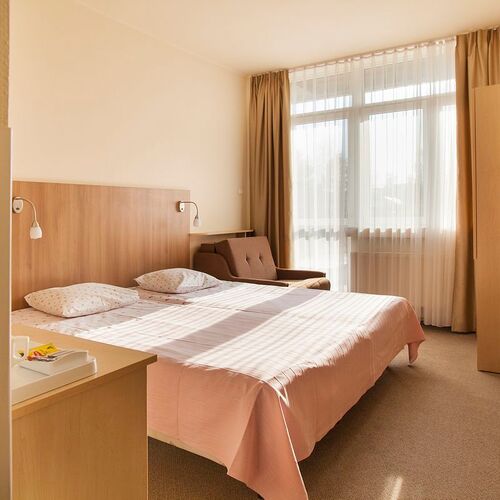 Hotel room with a double bed, wooden headboard, reading lamps, an armchair by a large window and a TV.