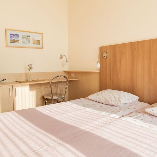 Bright, sunny room with a double bed and small floral bedding, a wooden headboard, a desk with a kettle, a chair and wall lamps.