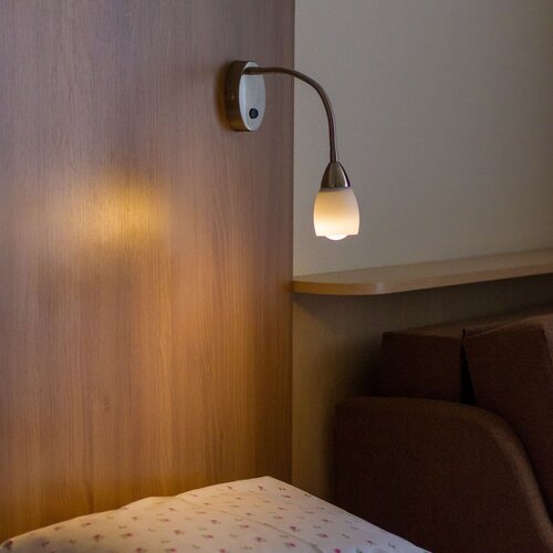 A bedside lamp above the bed with a wooden headboard, floral bedding and a cushion, a cozy corner with an armchair