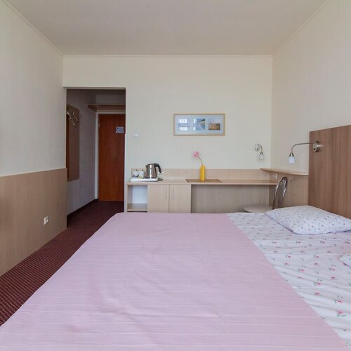 Hotel room with a bed in a pink throw, floral bedding, a wooden headboard, TV, desk and kettle.