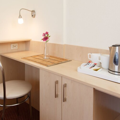 Desk in a hotel room with an electric kettle, cups, a lamp, a chair and a vase with pink flowers