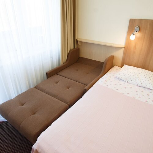 Hotel room with a double bed and a brown sofa bed, wooden headboard, lamp, light curtains.