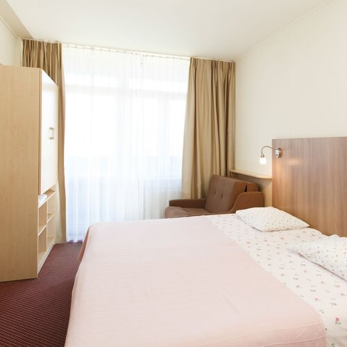 Bright hotel room with a large bed in pink bedding, a wooden headboard, wall lamps, a TV, wardrobe and an armchair.