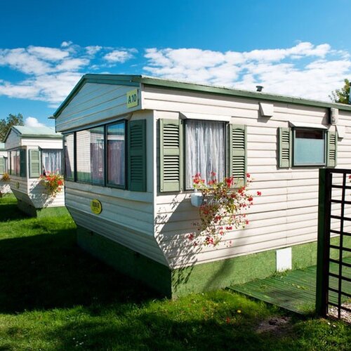 A row of light mobile homes with green shutters and flower pots on the lawn; the Eden Spa tower in the background.