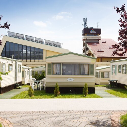 A complex of mobile homes with terraces and plastic garden furniture on the lawn, the resort with a tower and the 'eden' logo in the background.