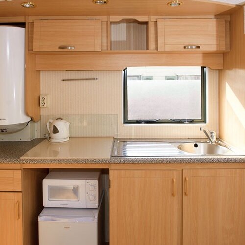 Compact kitchenette with light wooden cabinets, a sink by the window, a kettle, microwave and fridge.