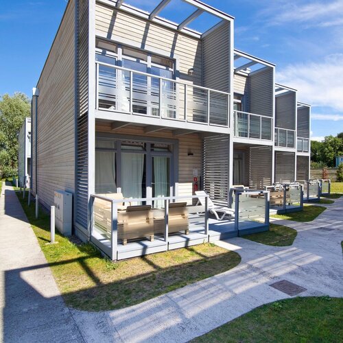 Modern rooms with a private terrace and upper balcony, wooden facade, glass balustrades, patio furniture and sunbeds on the lawn.