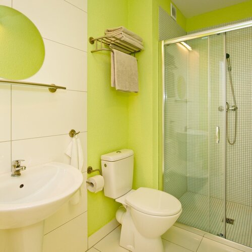 Hotel bathroom with green walls, a round mirror, a sink, a toilet, a glass shower enclosure and towels.