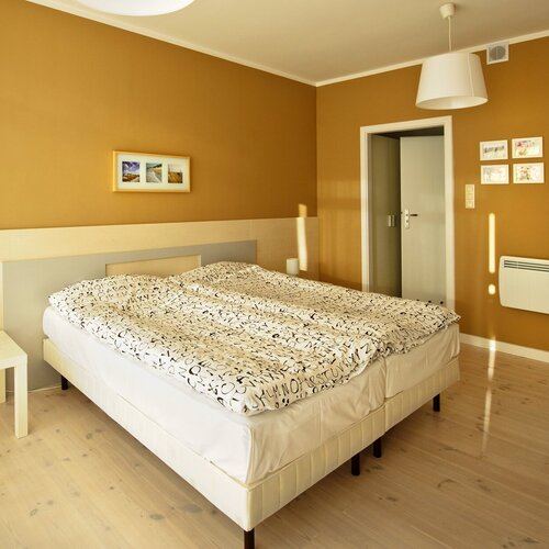 Cozy hotel room with a large double bed, light bedding with a letter pattern, a bedside table, a floor lamp and a warm yellow wall.