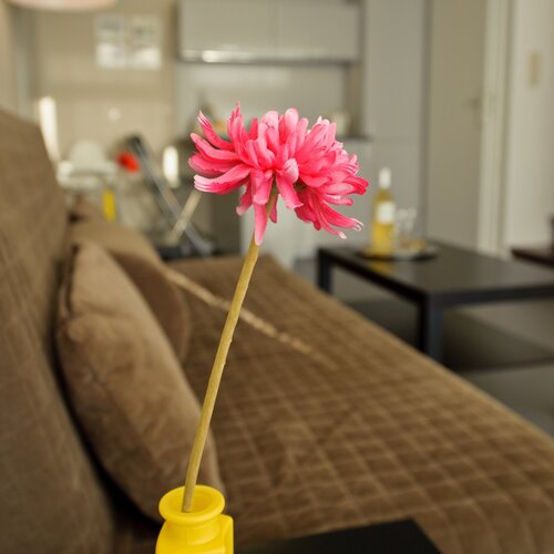 A pink flower in a yellow vase on a table in front of a brown sofa; a kitchenette and a bottle of wine in the background