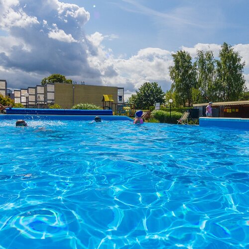 Sunlit outdoor hotel pool with bright blue water, guests swimming, deck chairs, lifebuoy, landscaped gardens and cloudy sky.