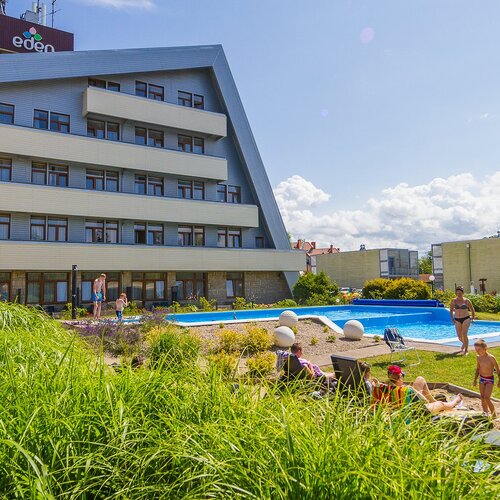 Hotel with a slanted roof and balconies, an outdoor pool with loungers, a lawn and families with children playing by the water
