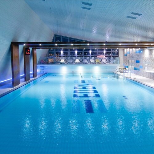 Indoor hotel pool with blue lighting, wooden beams, a sunbed and a spa area with a person resting.