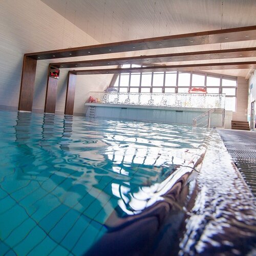 Indoor hotel pool with large windows, wooden beams, entry steps and calm blue water