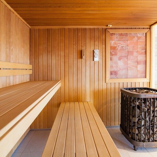 Wooden hotel sauna with two rows of benches, a stone heater, a decorative salt wall and a glazed entrance to the spa area