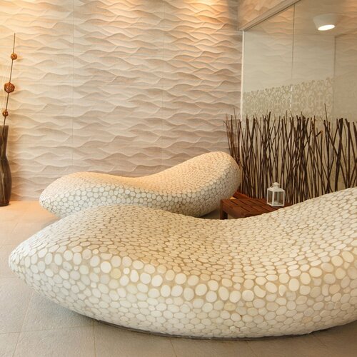 Spa relaxation area with two sculptural stone-mosaic loungers, a wavy wall, soft light, a vase and a glass partition.