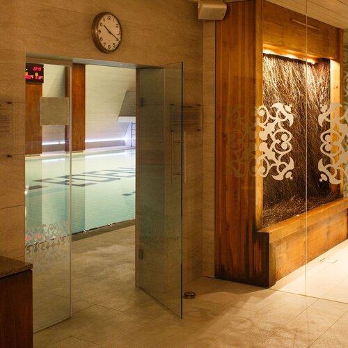 Indoor pool behind glass doors, decorative matte patterns, a wooden water wall, white benches and warm lighting.