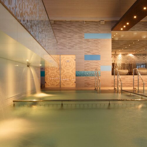 Indoor spa pool with water cascades, warm lighting, handrails and relaxing chairs.