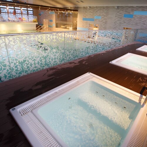 Spa area with three jacuzzis on a wooden terrace, decorative glass balustrades and a view of the indoor pool