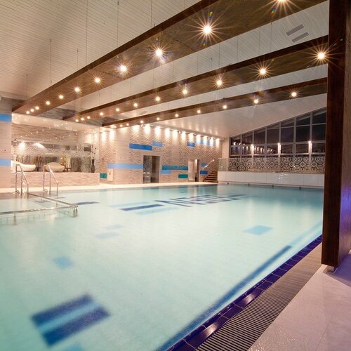 Large indoor hotel pool with illuminated water, wooden ceiling beams, sunbeds and a glazed viewing wall