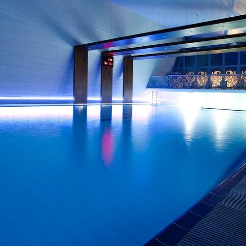 Indoor hotel pool with calm, blue-lit water, wooden beams and a terrace with loungers.