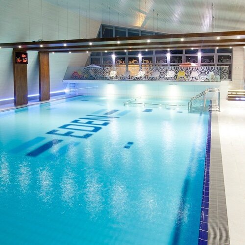 Elegant indoor hotel pool with turquoise water, lighting, a spacious terrace, steps and a view of a decorative glazed gallery.