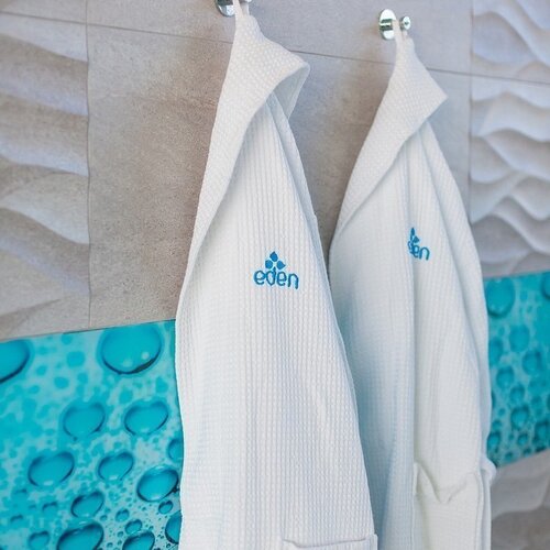 White spa robes with embroidered Eden logo hanging on chrome hooks against a backdrop of wavy tiles and a turquoise panel