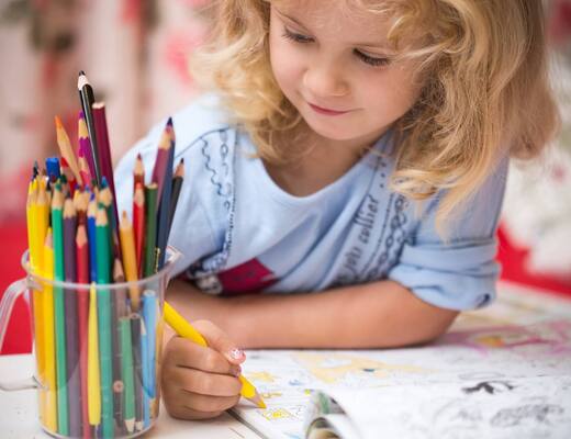 A girl coloring in a book, holding a yellow crayon next to a clear cup with colorful crayons — the hotel's family play corner.