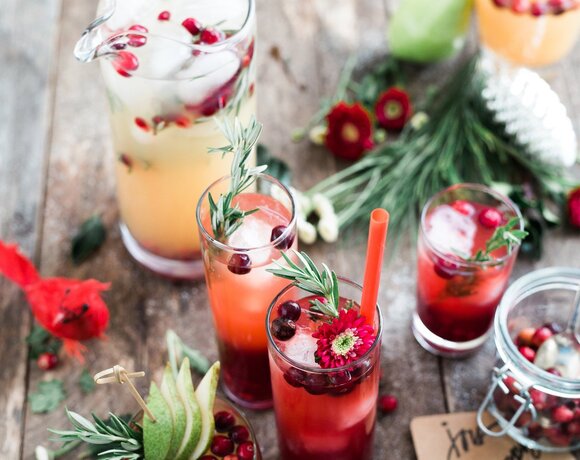 Cocktails and a jug of fruit lemonade on a rustic table, garnished with cranberries, rosemary, pear slices and a flower.