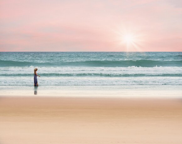 A woman in a dress by the edge of a sandy beach, gentle waves and a pink sky at sunset