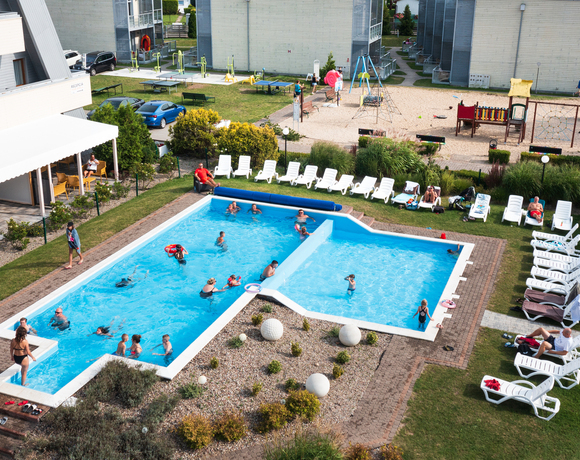 Hotelowy basen zewnętrzny z rodzinami pływającymi, rzędem leżaków, zadaszoną strefą wypoczynku i placem zabaw w tle