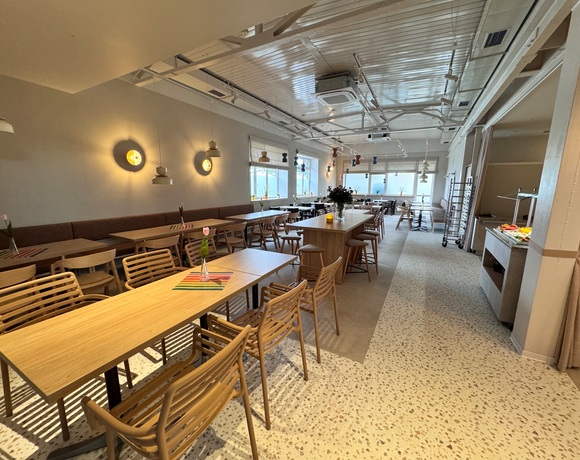 Przytulna jadalnia śniadaniowa z drewnianymi stolikami, centralnym stołem barowym, podwieszanymi lampami i bufetem z owocami.