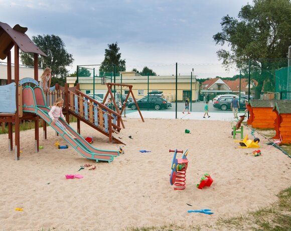 Rodzinny, ogrodzony plac zabaw na piasku przy hotelu z drewnianą zjeżdżalnią, bujakami i boiskiem w tle.