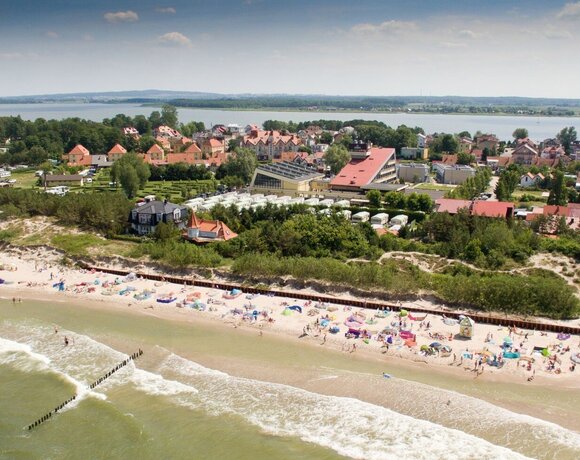 Beach with a sandy shore and colorful umbrellas, a waterfront hotel complex with red roofs, dunes, wooden groynes and a bay in the background