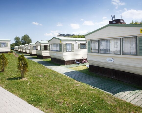 Rząd mobilnych domków letniskowych przy drewnianych ścieżkach, zielony trawnik, zasłonięte okna i dziecięce rowerki przy wejściach