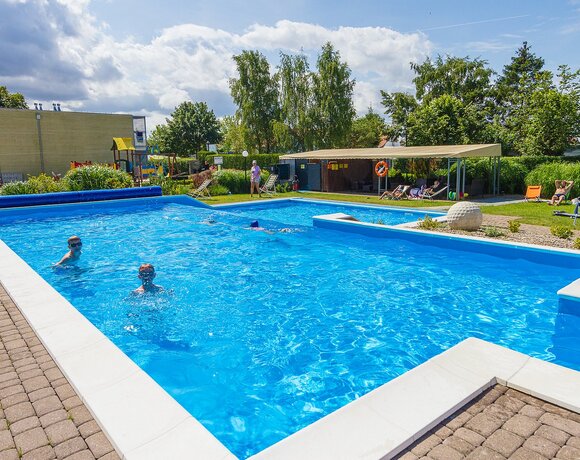 Hotelowy zewnętrzny basen z krystalicznie niebieską wodą, leżakami, zadaszonym patio, placem zabaw i kąpiącymi się dziećmi.