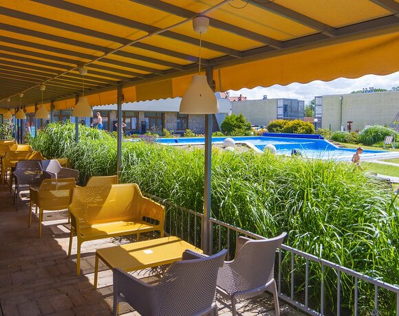 Zadaszony taras z żółtymi markizami i miejscami do siedzenia, widok na basen, zieleń i bawiące się dziecko.