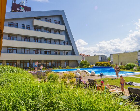 Hotel ze skośnym dachem i balkonami, zewnętrzny basen z leżakami, trawnik i rodziny z dziećmi bawiące się przy wodzie