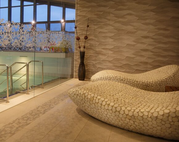 Relaksacyjny kącik spa z dwiema falistymi leżankami w kamiennym wzorze, przeszklony basen i teksturowana ściana.