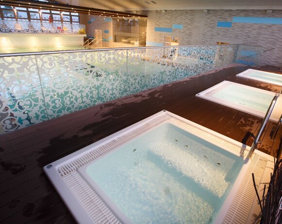 Strefa spa z trzema jacuzzi na drewnianym tarasie, ozdobne szklane balustrady i widok na basen wewnętrzny