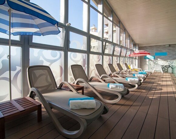 Przeszklona strefa relaksu z rzędem nowoczesnych leżaków, zwiniętymi ręcznikami, parasolami i drewnianą podłogą.