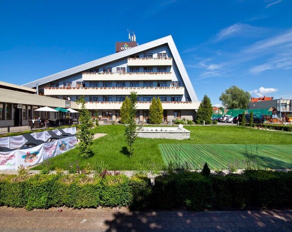 Hotel z skośnym dachem i balkonami z kwiatami, zadbany trawnik z leżakami, taras z parasolami i plac zabaw