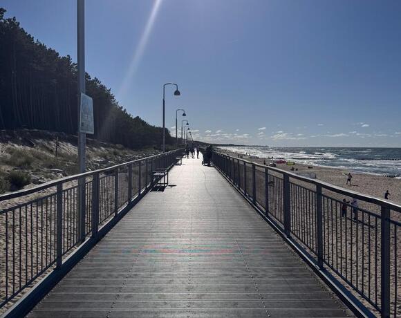 A sunny seaside promenade with lamps and benches, a pine forest to the left and waves breaking on the sandy beach