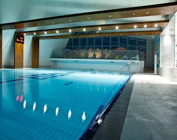 Spacious illuminated indoor pool with wooden beams, a gallery with loungers and stairs with a handrail.