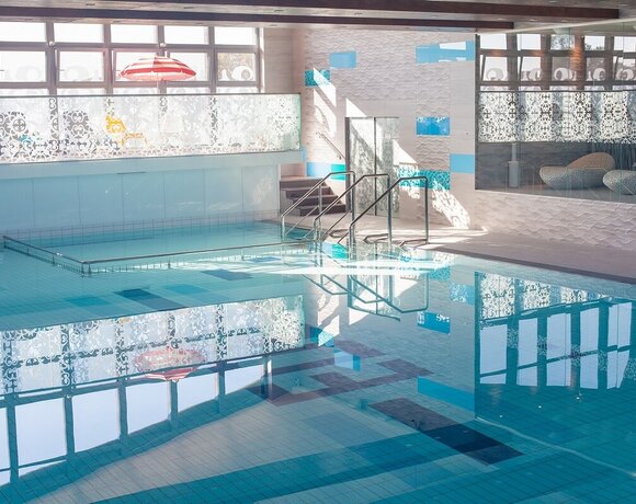 Indoor hotel pool with turquoise water, steps and handrails, decorative partitions and a bright relaxation area