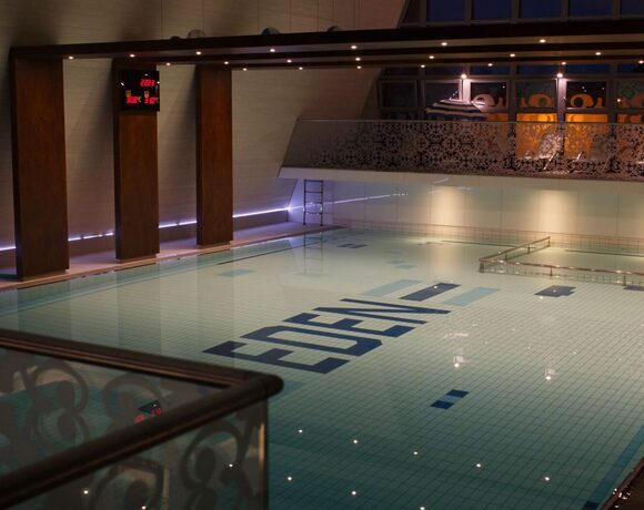 Indoor heated hotel pool with mosaic tiles, a decorative balcony with loungers, handrails and atmospheric lighting